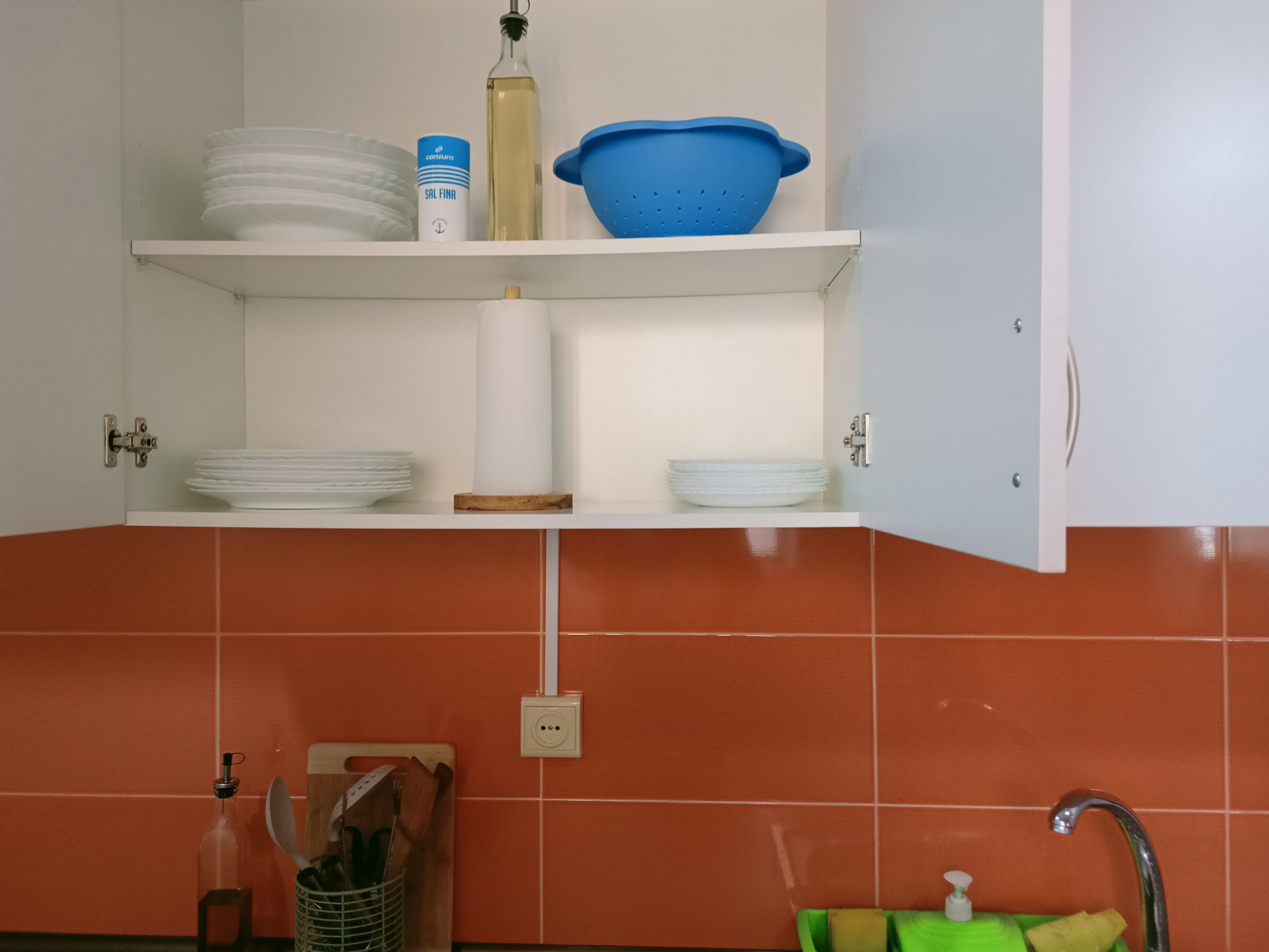Ett kök med vita hyllor där det står tallrikar, en flaska och ett blått durkslag.