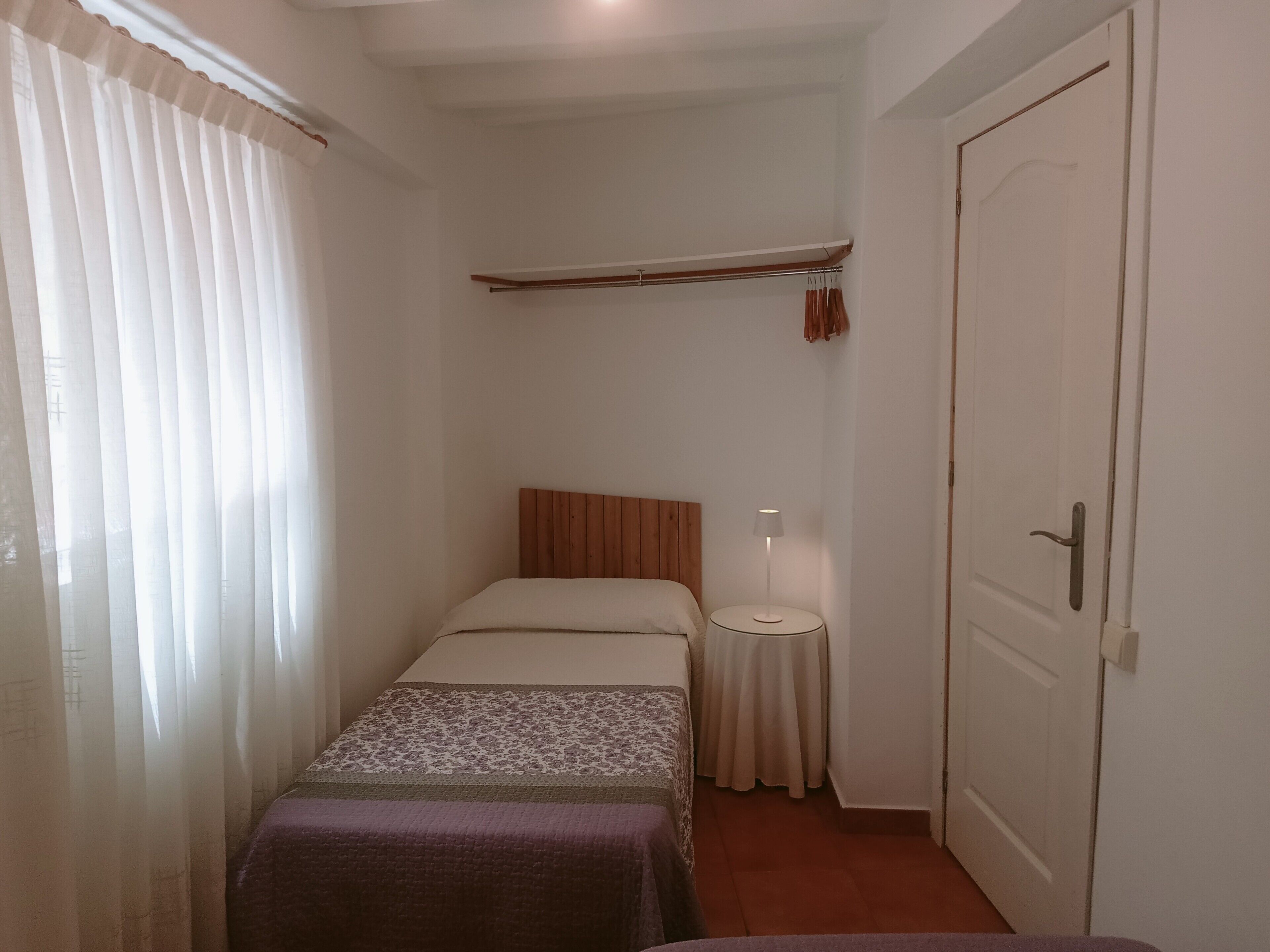 Una habitación pequeña y estrecha con una cama individual, una mesita de noche con lámpara, un perchero y una puerta.