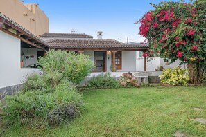 Una casa con techo de tejas, puerta principal y un pequeño jardín con diversas plantas y flores.