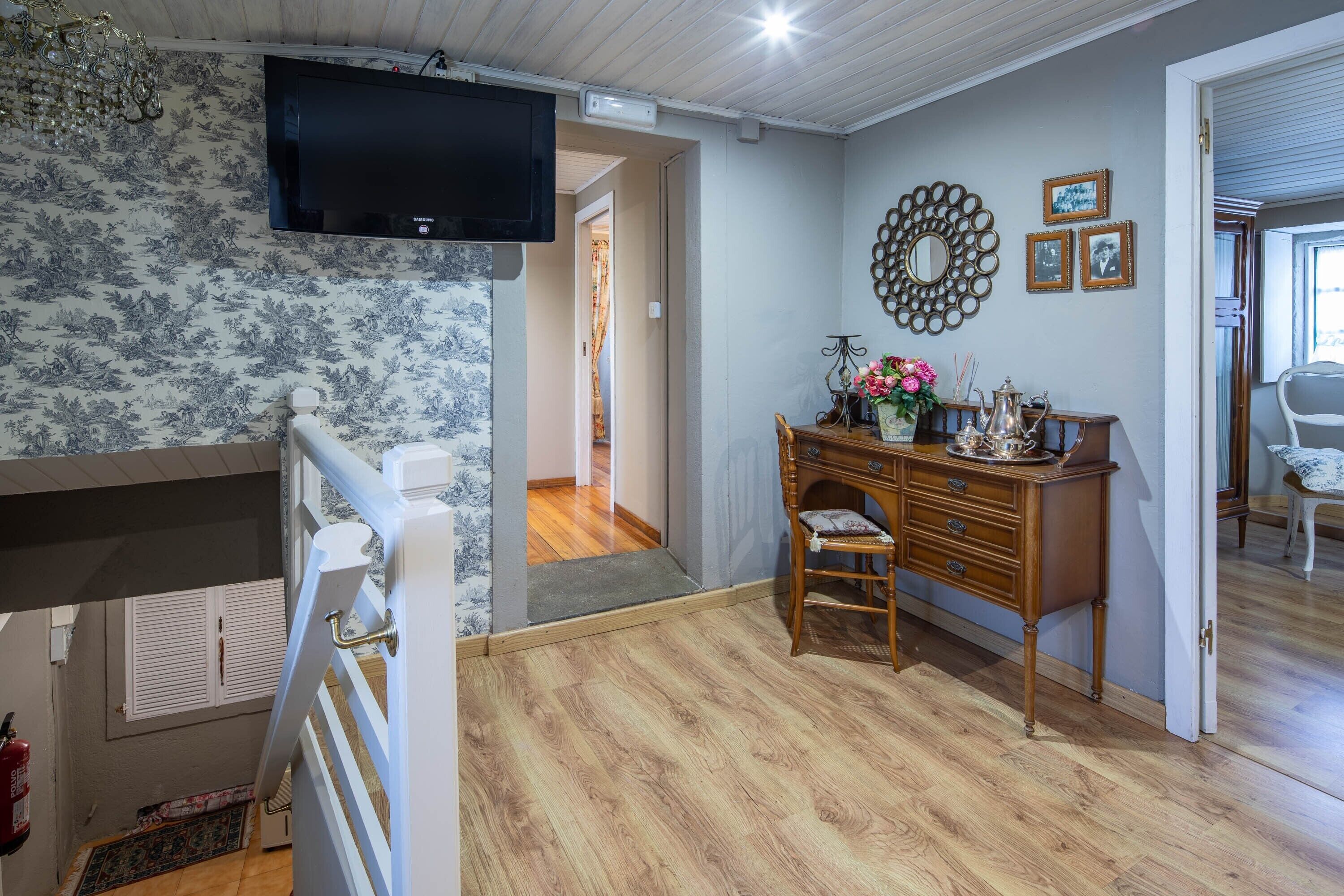 A room with a wooden floor, a desk with drawers, a chair, a television mounted on the wall, and a door leading to another room.