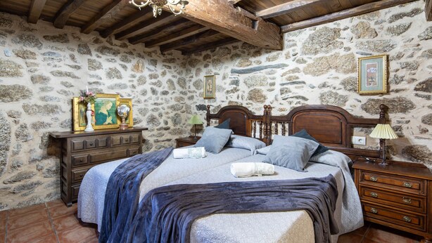 A bedroom with a stone wall, wooden bed frame, and a nightstand with a lamp.