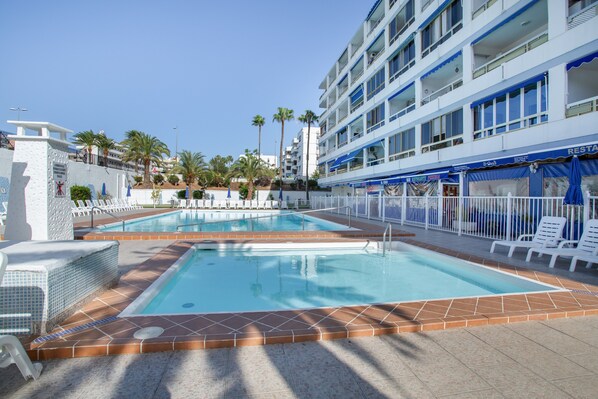 Zona de piscina hotelera con dos piscinas, tumbonas y un edificio con balcones.