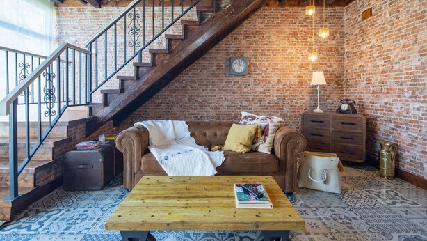 A cozy room with a wooden staircase, a brown leather sofa, a wooden coffee table, a lamp, and a clock on the brick wall.