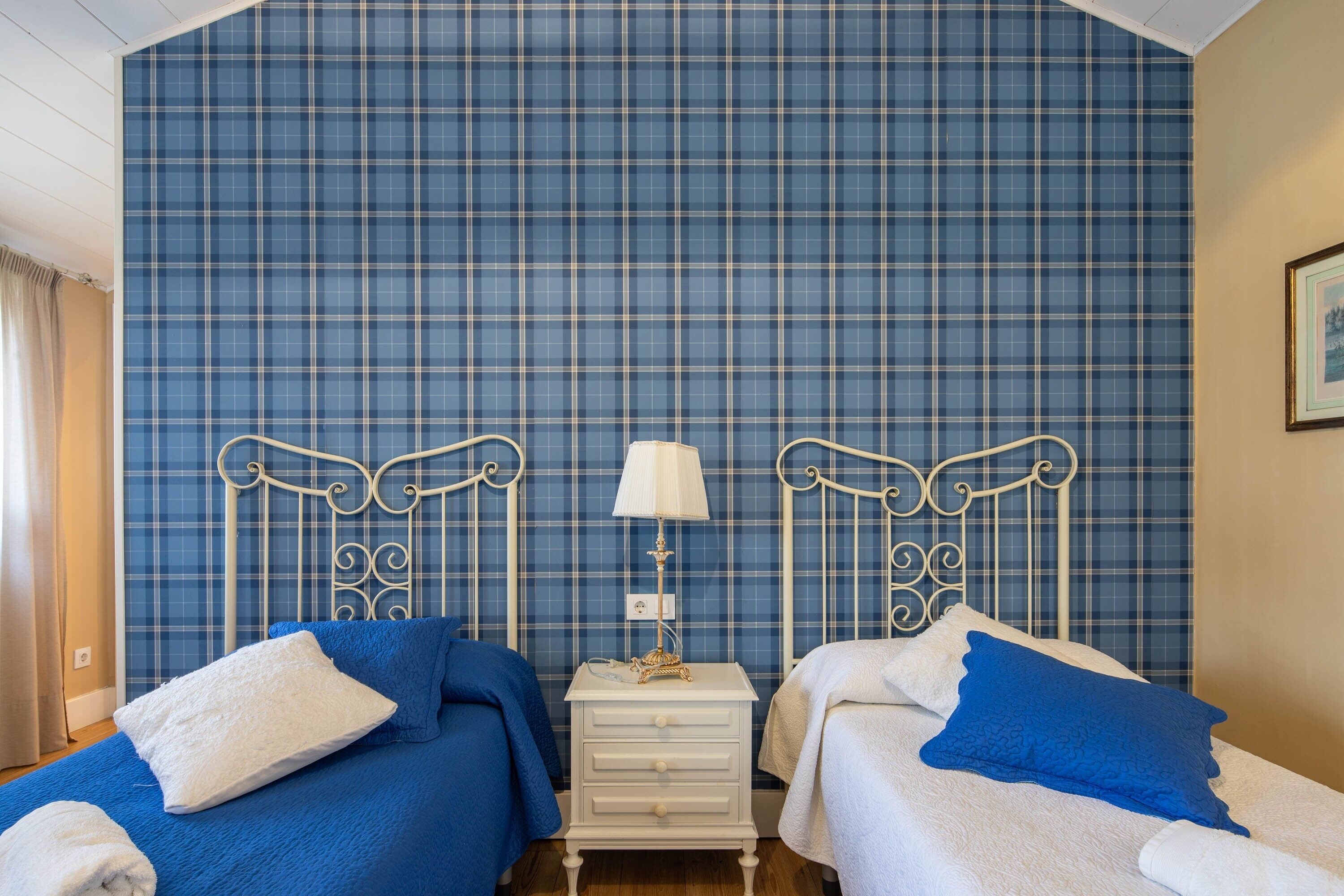 Una habitación con dos camas, una mesita de noche con lámpara y papel tapiz azul a cuadros.