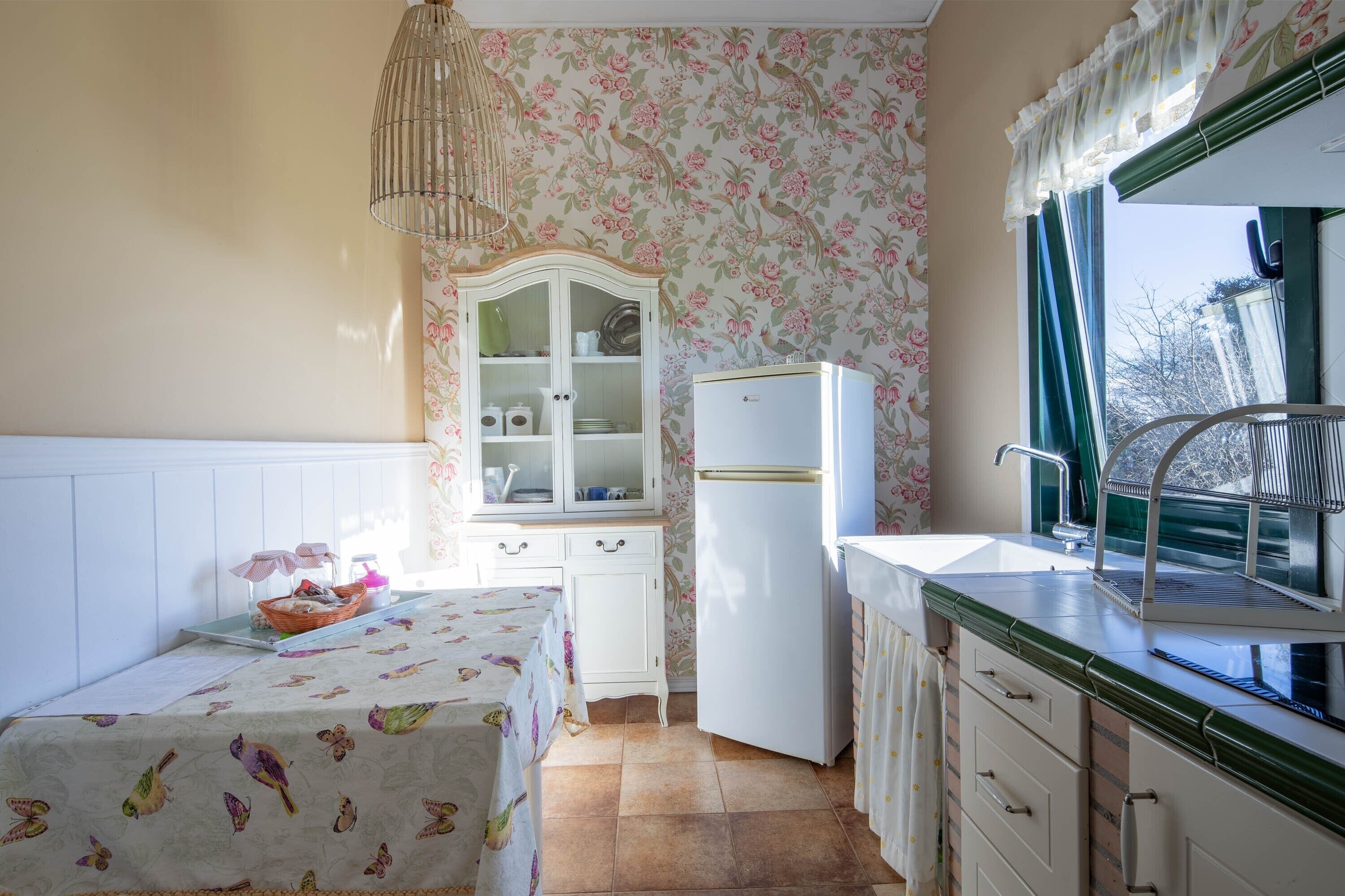 Una cocina con un refrigerador blanco, un lavamanos con ventana, un armario y una mesa con mantel floral.