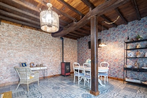 Une pièce rustique avec des poutres apparentes, un mur en briques, des carreaux de sol à motifs, une table à manger avec des chaises, un fauteuil en osier, une étagère avec des objets décoratifs et un luminaire suspendu.