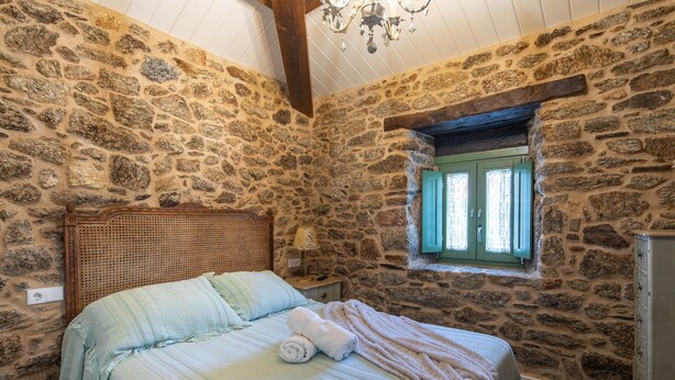 Un dormitorio con paredes de piedra, una cama, cabecero de madera, mesita de noche, lámpara y ventana con contraventanas.