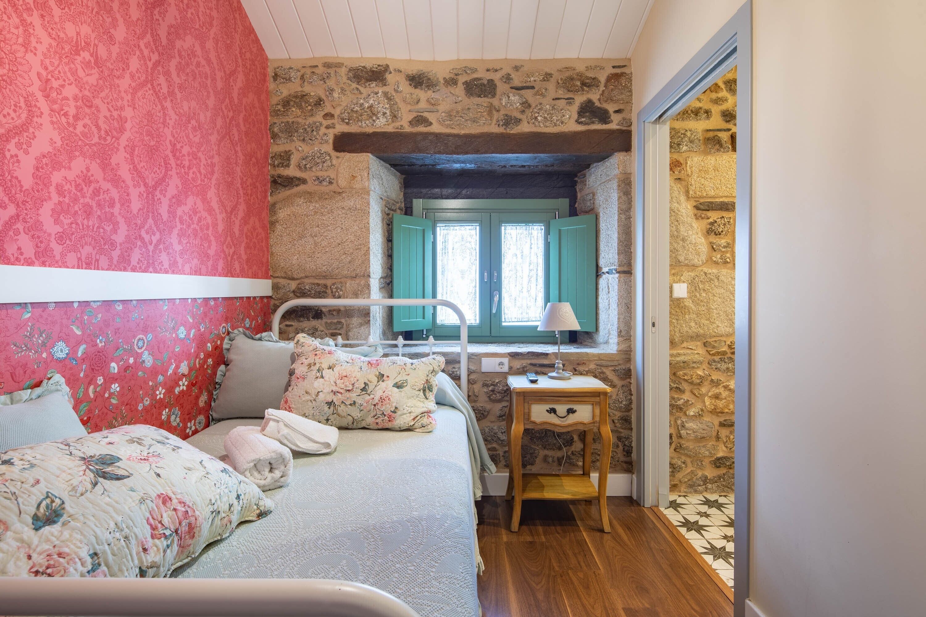A bedroom with a stone wall, a bed with floral bedding, a bedside table with a lamp, and a window with green shutters.