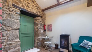 Una habitación de paredes de piedra con una puerta verde, una pequeña mesa con una silla, un sillón verde y un estufa de leña.