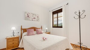 A bedroom with a bed, nightstands, a window with bars, a decorative metal pole, and a floral artwork on the wall.