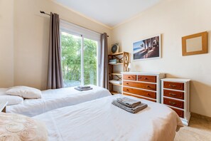 Quarto de hotel com duas camas, uma cômoda, uma estante com um ventilador e uma janela com cortinas.