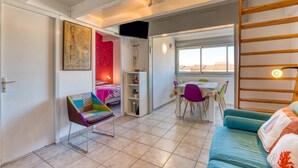A room with a bed, a chair, a table, and a sofa. There is a window with a view of buildings outside. - Apartment 'La Conque' with Air Conditioning (Agde)