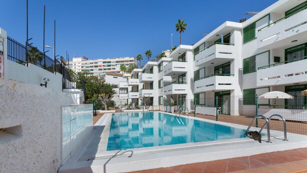 A swimming pool with a tiled deck, surrounded by multi-story buildings with balconies and palm trees.