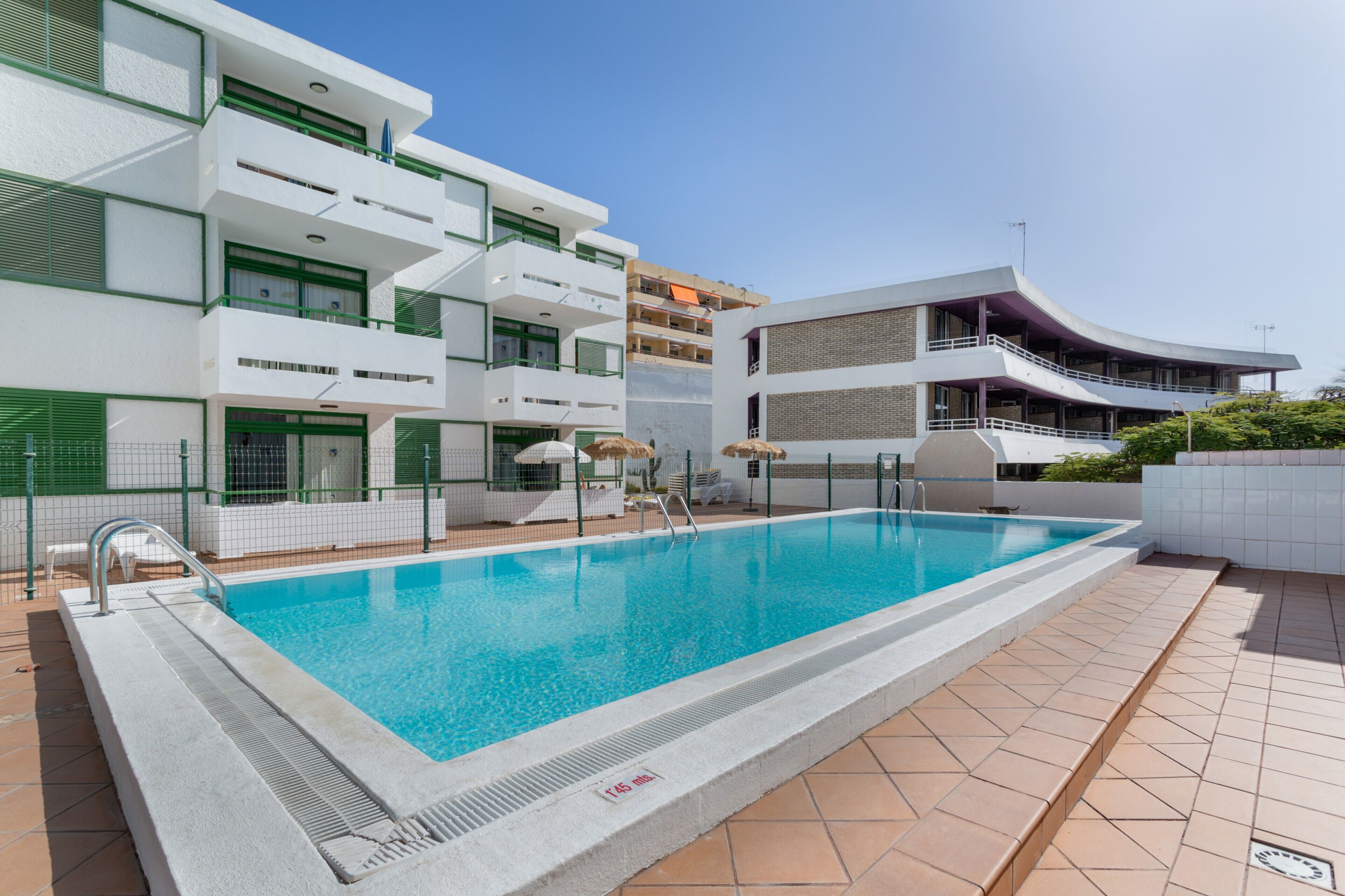 Una piscina con solárium y azulejos, un edificio blanco con balcones y una estructura curva también con balcón.