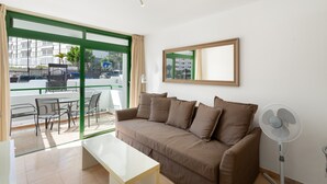 A living room with a sofa, a table, a fan, and a balcony with a view of buildings.