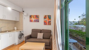 A compact living space with a sofa, a small table, a washing machine, and a window with a view of outdoor seating.