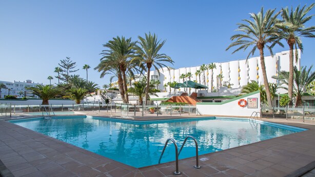 A swimming pool with a tiled deck, palm trees, and a building with balconies in the background.