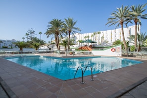 Une piscine avec une terrasse carrelée, des palmiers et un bâtiment doté de balcons en arrière-plan.