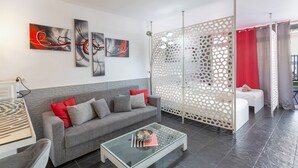 Un salón moderno con un sofá gris, cojines rojos, arte abstracto en la pared, una mesa de centro de vidrio y un separador blanco con diseño perforado.