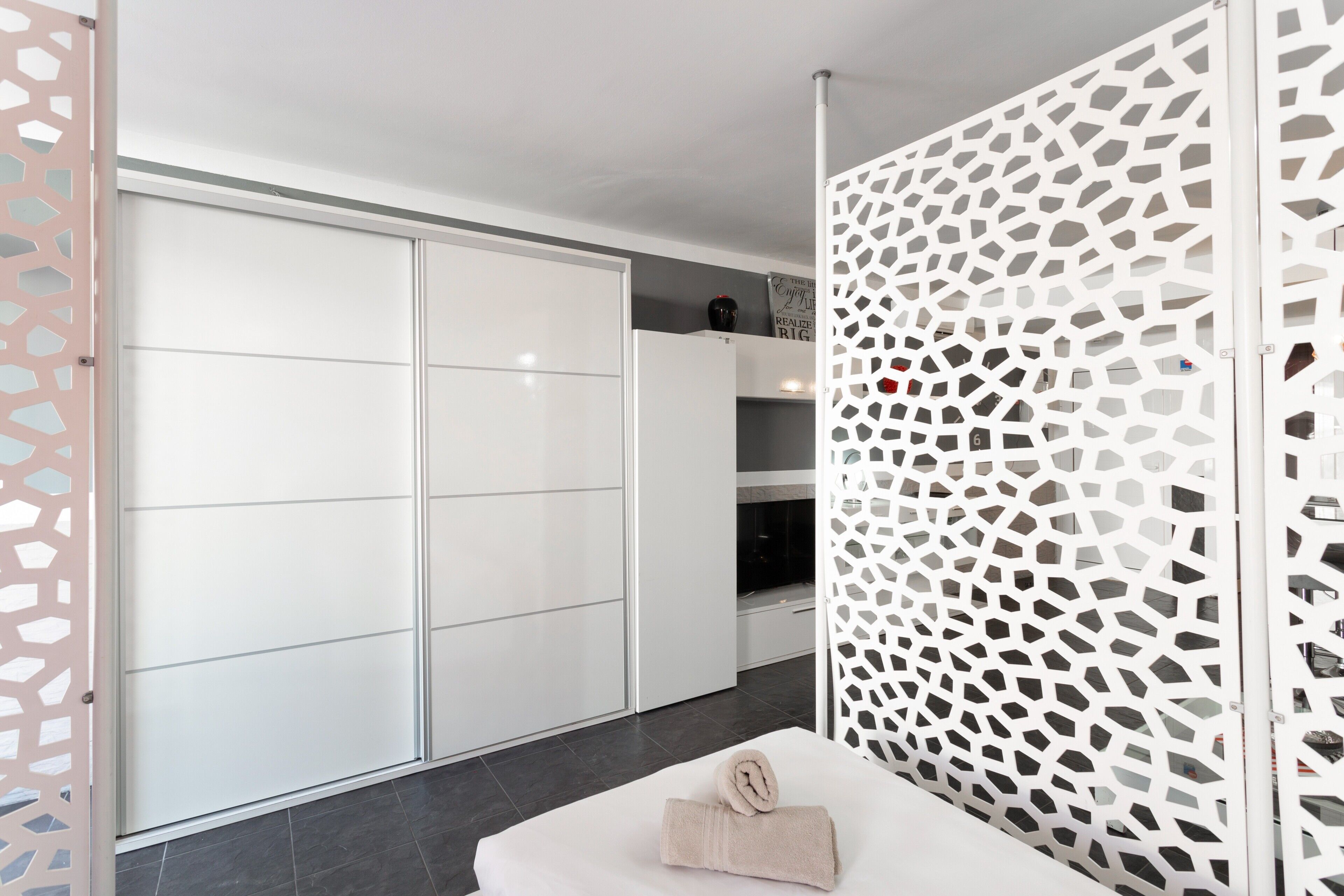 Une chambre moderne avec une armoire blanche, un panneau décoratif blanc et un lit bien fait, sur lequel est posée une serviette enroulée.