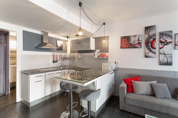 A modern kitchen with a central island, bar stools, and a grey sofa with red cushions.