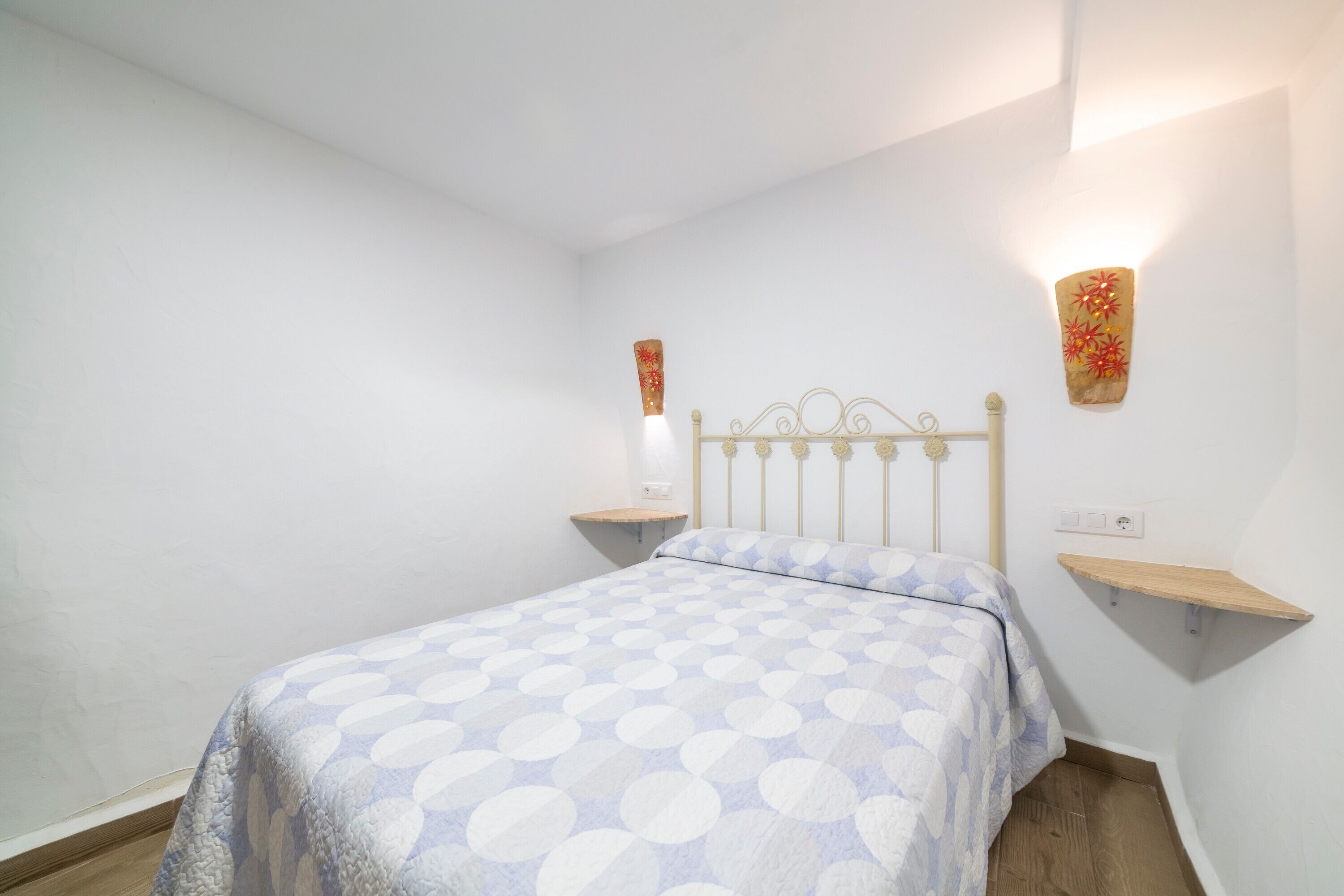 A bedroom with a bed, a nightstand, a wall-mounted light, and a shelf.