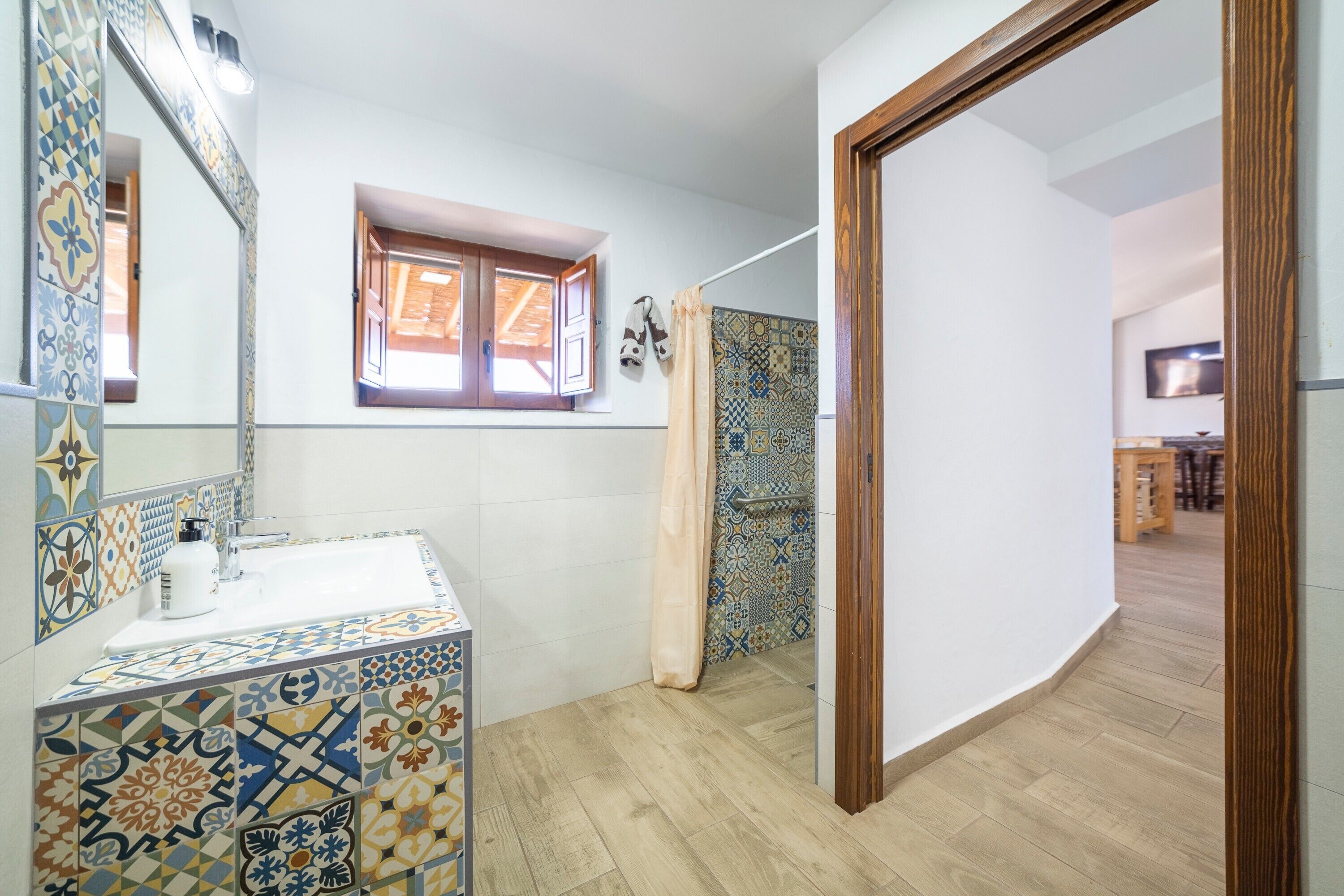 A bathroom with a tiled floor, a sink with a mirror, a window with wooden shutters, and a shower curtain.