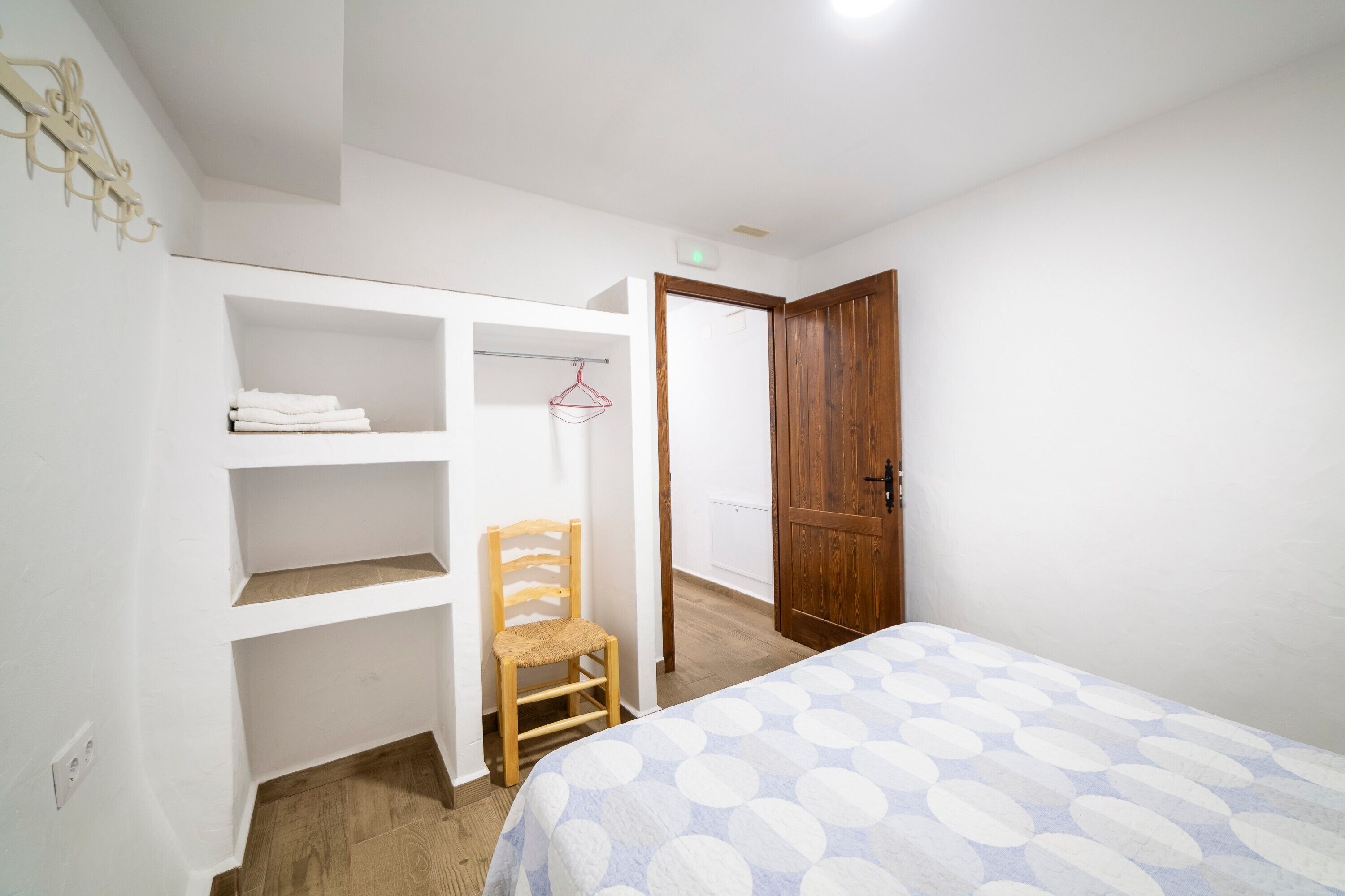 Un dormitorio con cama, una silla de madera, un armario y una puerta que da a otra habitación.