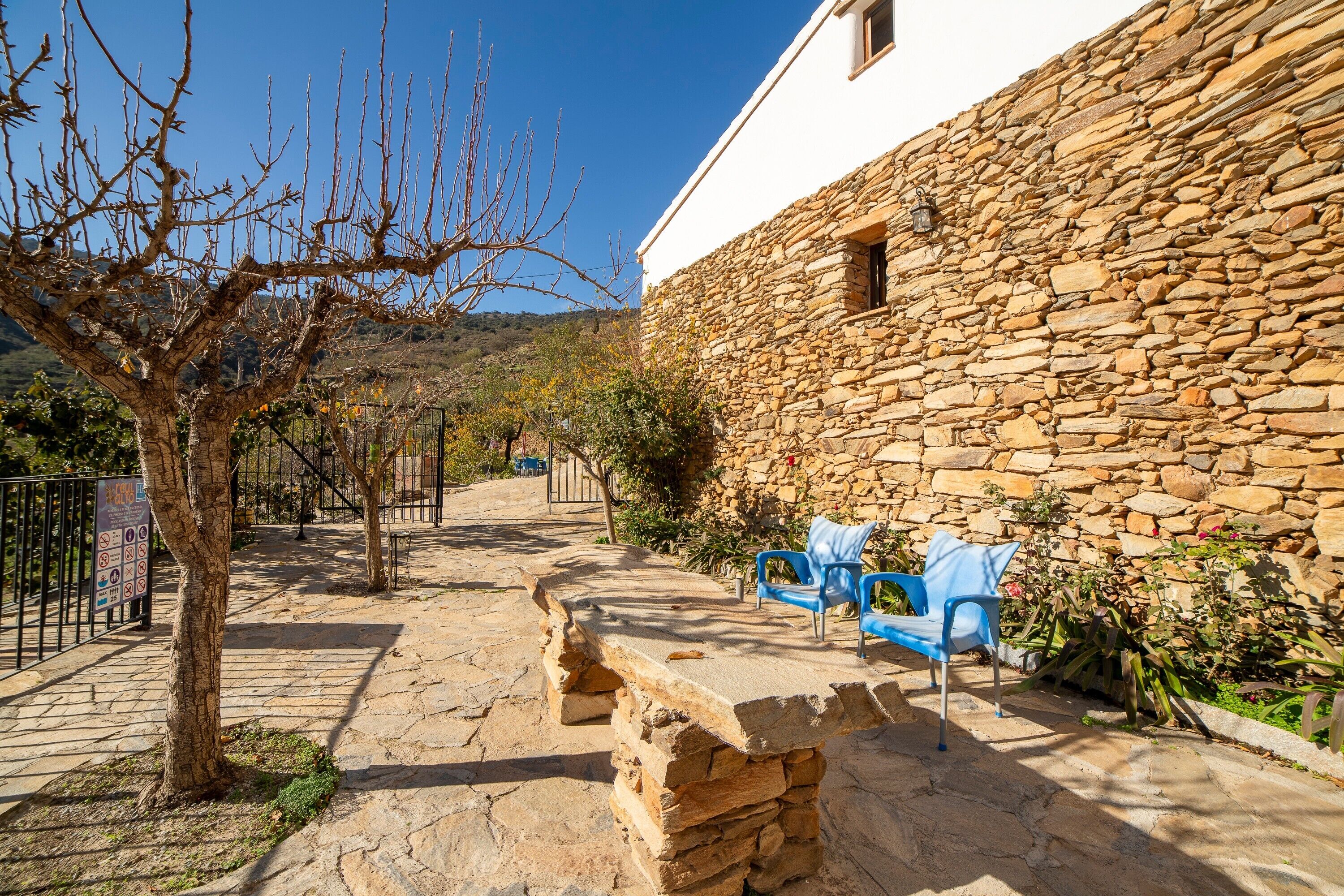 A stone building with a stone wall, a tree, and two blue chairs.