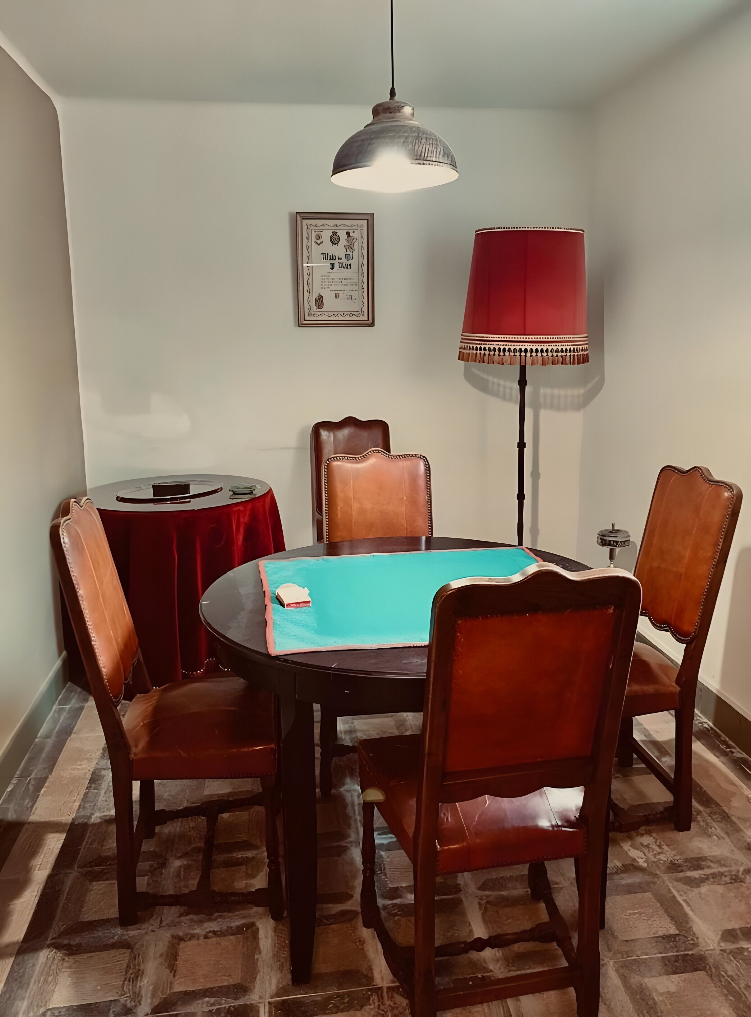 A dining area with a round table, four chairs, a red tablecloth, a pendant light, and a framed picture on the wall.