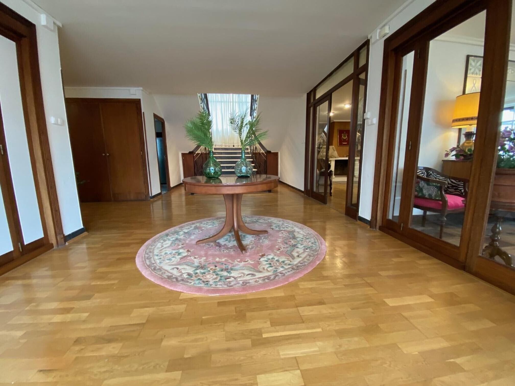 Un pasillo amplio con una mesa redonda en el centro, flanqueado por puertas de madera y vistas a una habitación con una silla y una lámpara.