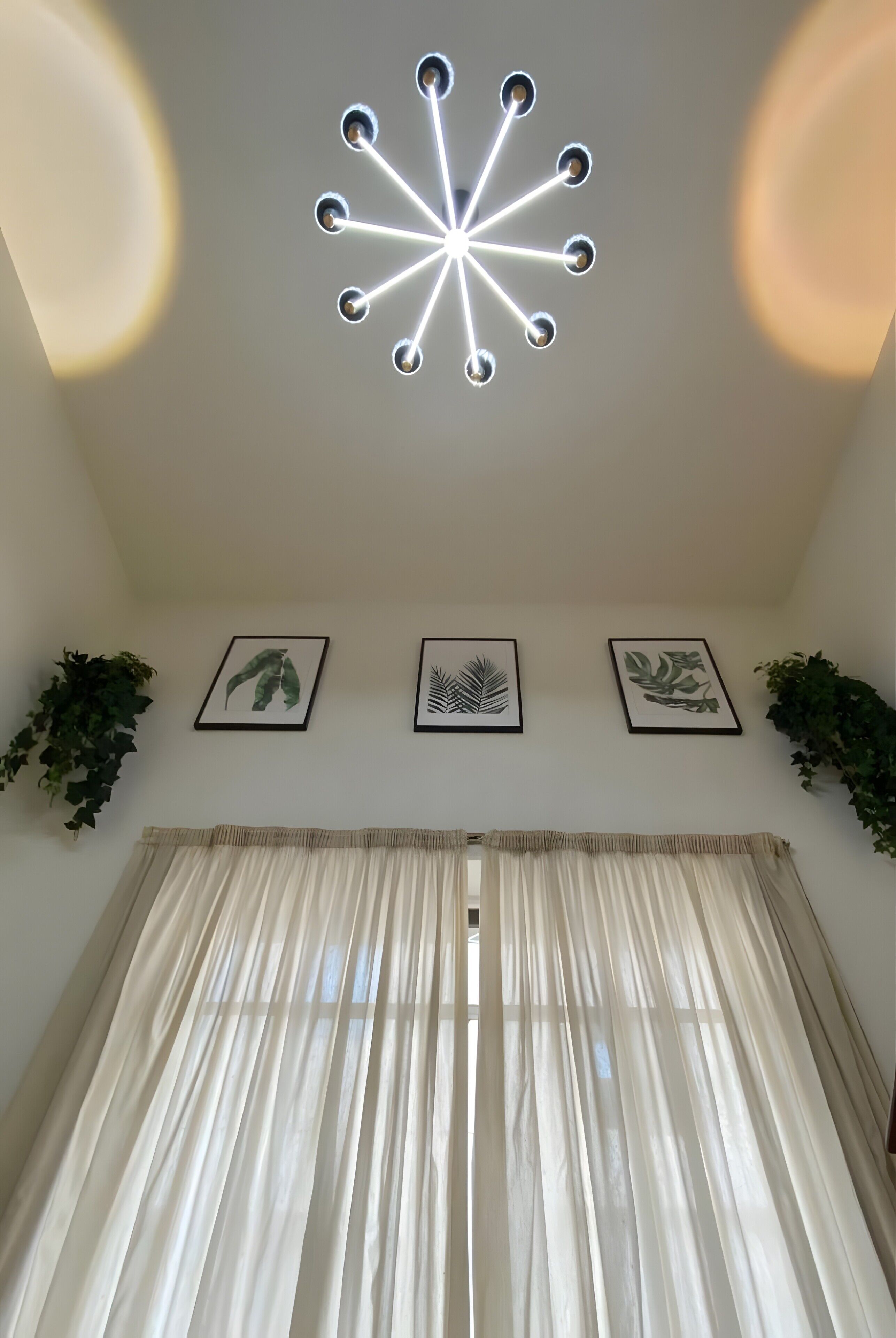 Una habitación con una lámpara en el techo, tres cuadros en la pared y una ventana con cortinas traslúcidas.