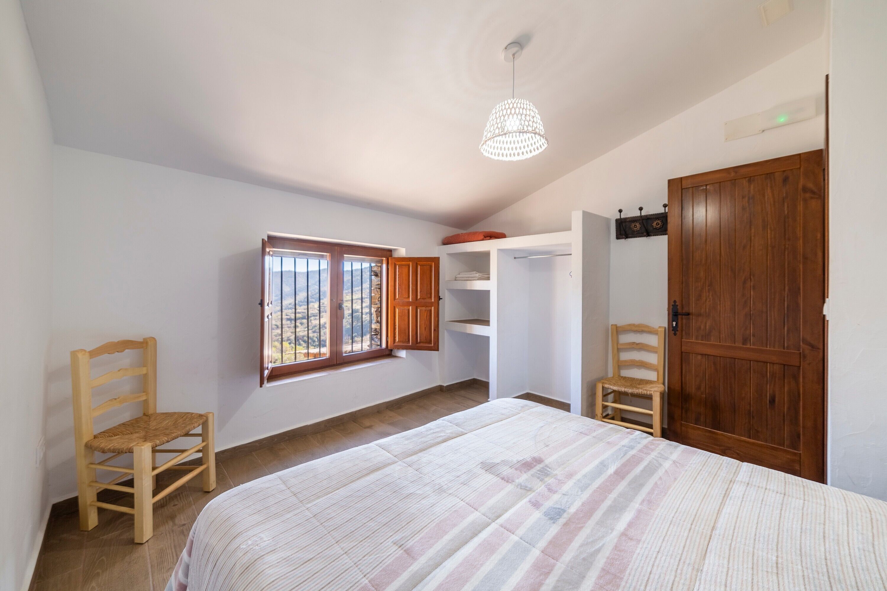A bedroom with a bed, two wooden chairs, a wardrobe, a window with a view, and a hanging light fixture.