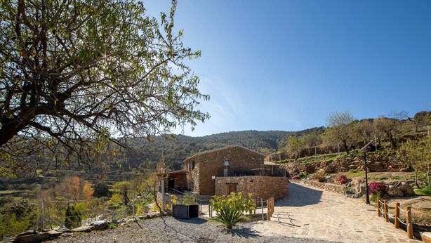 A rustic stone building with a balcony, surrounded by a gravel path and greenery.