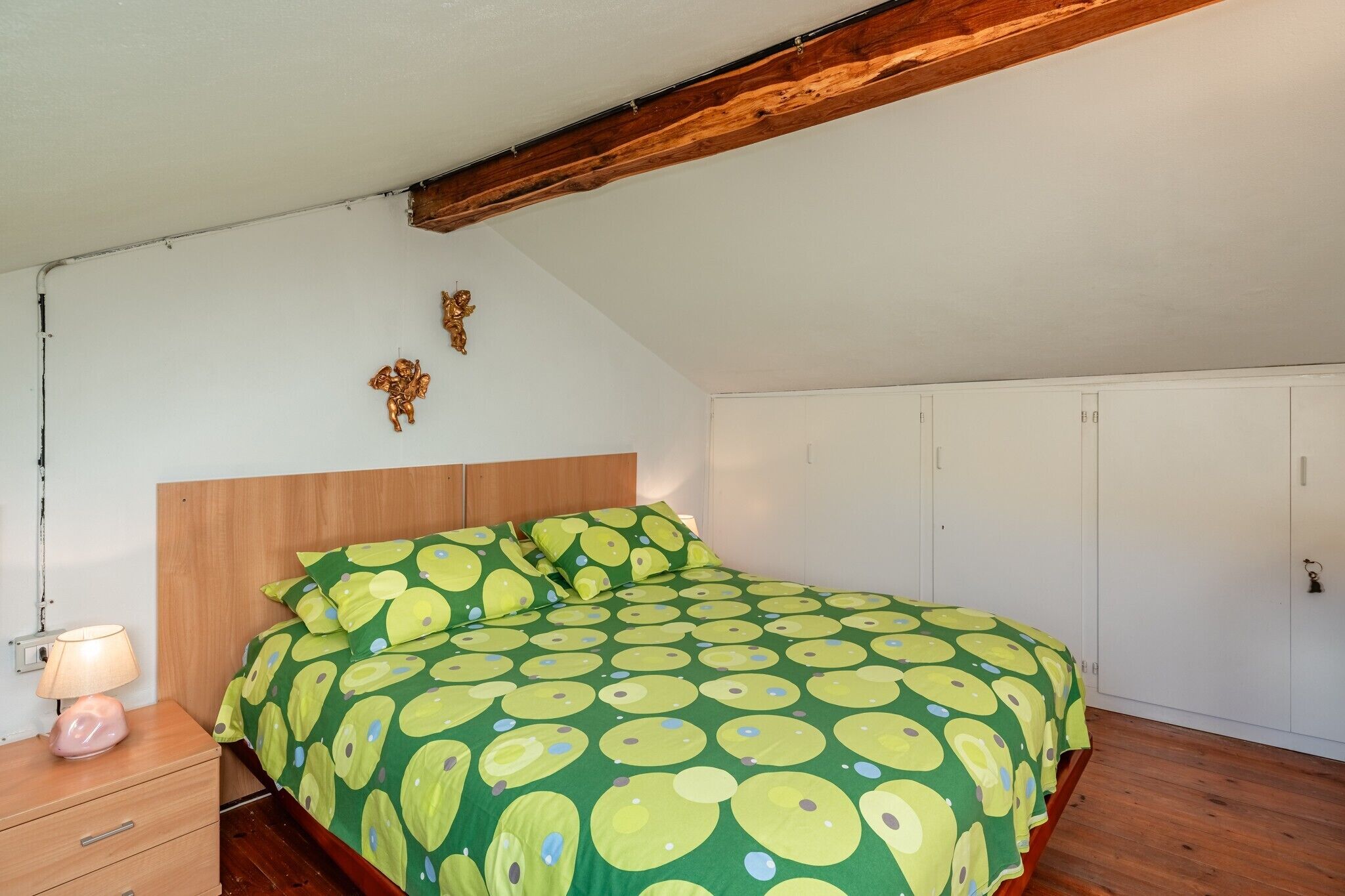 Un dormitorio con cabecera de madera, cama con colcha de motivos aguacate en verde y amarillo, lámpara de noche y armario blanco.