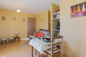 A dining area with a table set for two, a bookshelf, and a door in the background. - Holiday Home 'Gîte Proche Mer 800 M' with Private Terrace, Shared Garden and Wi-Fi (Plouézec)