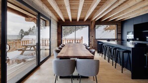 Une cuisine moderne avec une longue table en bois et des tabourets de bar, de grandes fenêtres donnant sur un paysage enneigé et un plafond en bois.