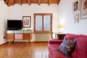 A room with a wooden floor, a red sofa, a television, a cabinet, a window with lace curtains, and wall art. - Apartment 'Casa Pòlse' with Mountain View, Shared Garden and Wi-Fi (Baselga di Pinè)