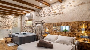Habitación con pared de piedra, vigas de madera, cama con ropa blanca, jacuzzi y panel de madera.