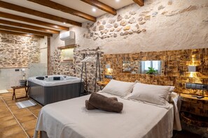 A room with a stone wall, wooden beams, a bed with white linens, a hot tub, and a wooden wall panel.