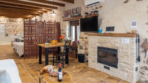 Un interior rústico con chimenea de piedra, vigas de madera, una zona de comedor con mesa y sillas, y un televisor montado en la pared.