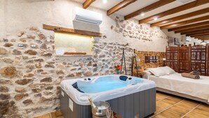 Una habitación de paredes de piedra con un jacuzzi, vigas de madera, una cama y una mesa de madera con un vaso y un cubo.