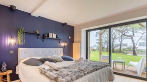 Une chambre avec un grand lit, une table de chevet, une plante suspendue, une étagère remplie de livres et une vue sur l’extérieur grâce à une porte vitrée.