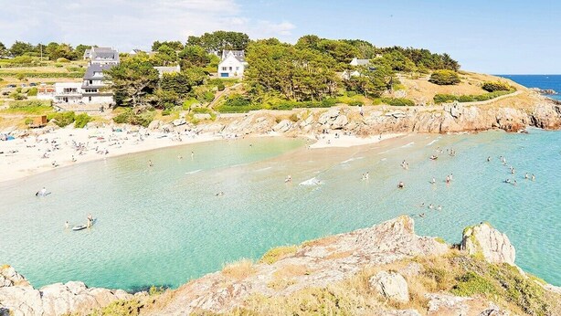 Une plage côtière aux eaux turquoise et limpides, avec un rivage rocheux et des bâtiments construits sur une colline.