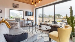 Un salon moderne comprenant un canapé, un fauteuil et une table basse, un téléviseur et une grande fenêtre donnant sur des immeubles et des arbres.