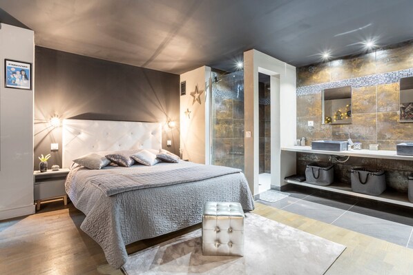 A modern bedroom with a large bed, bedside lamps, a nightstand, and a bathroom with a shower, mirror, and storage.