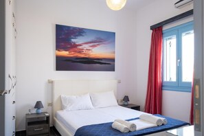 Ein Hotelzimmer mit Bett, Nachttischen, einem Sonnenuntergangsbild, einem Fenster mit roten Vorhängen und einer Klimaanlage.
