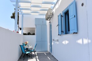 Um pátio externo estreito com uma mesa e cadeiras, uma parede branca com persianas azuis e uma porta.