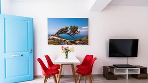 Une pièce avec une porte bleue, une table blanche et des chaises rouges, un téléviseur posé sur un meuble en bois, et un tableau représentant un paysage accroché au mur.