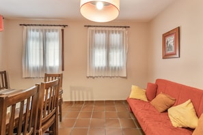 A room with a dining table, chairs, a sofa with cushions, and two windows with curtains.